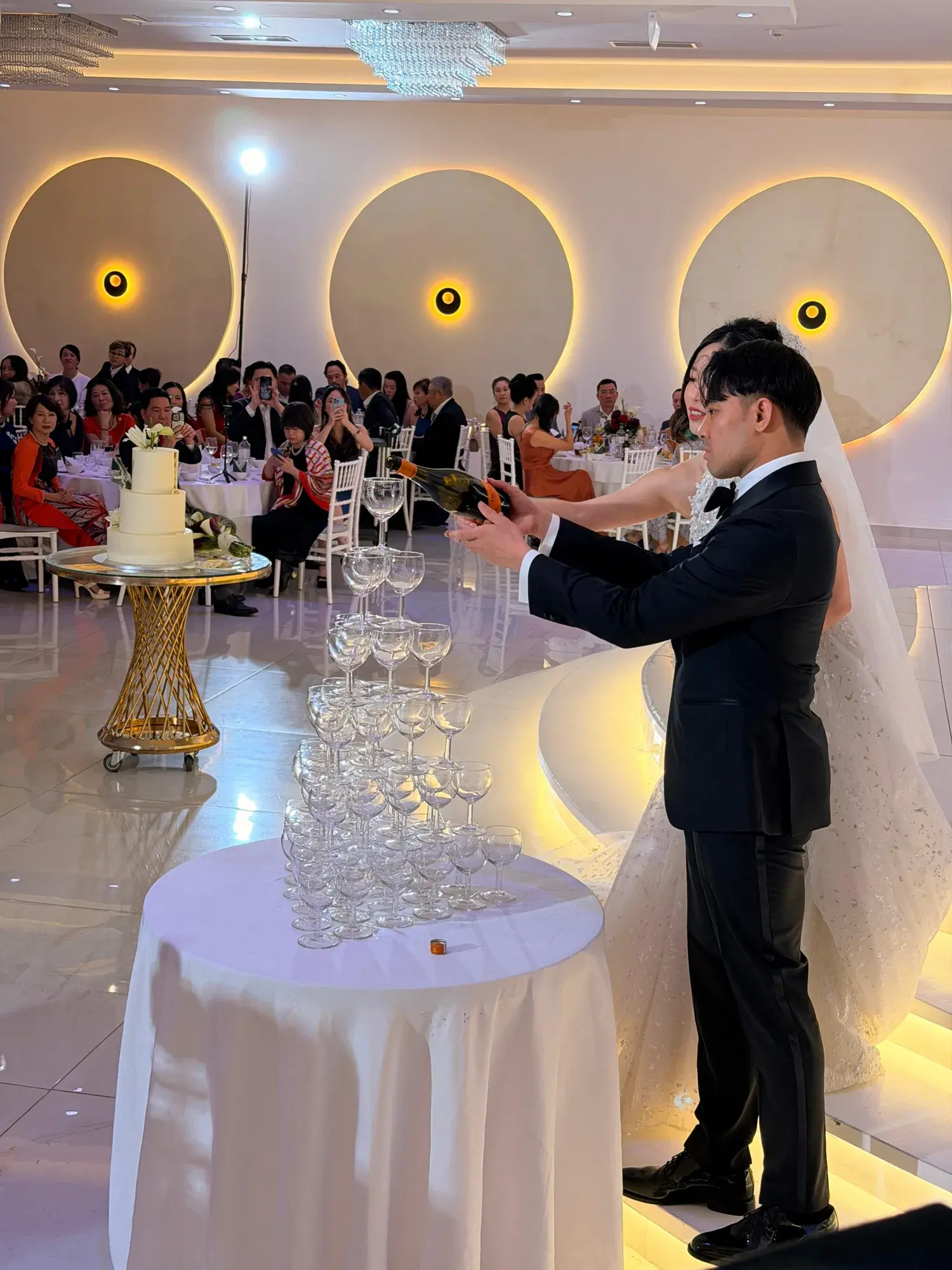 Brautpaar baut die Sektpyramide im weißen Saal des Moon Events, dahinter die beleuchteten Rundnischen, die Torte und die Gäste an den Tischen