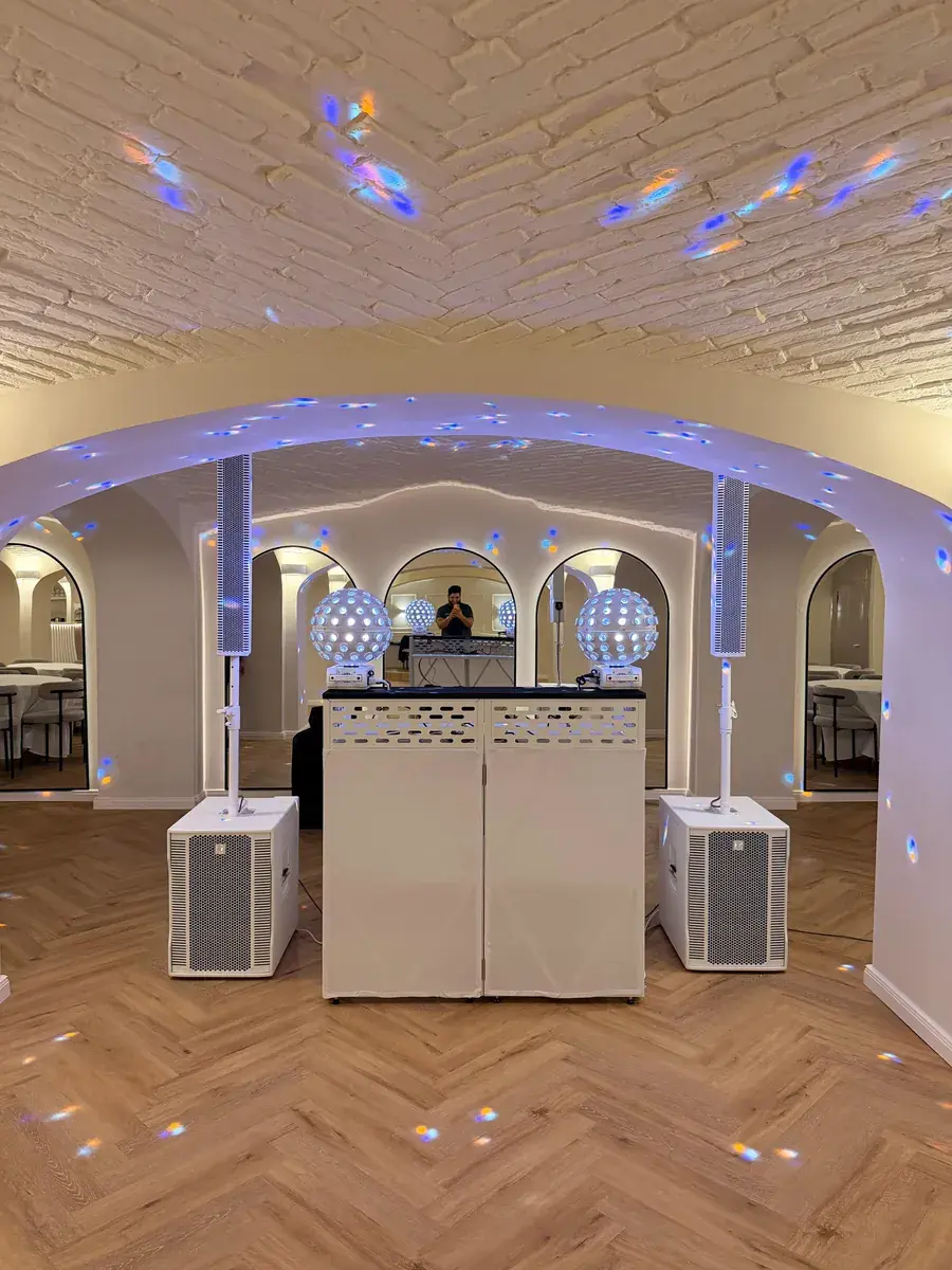 Weißes DJ-Kido-Setup mit DJ-Pult, zwei Säulenlautsprechern auf Subwoofern und Kugel-Lichteffekten im hellen Gewölbesaal von PERA Events unter einer Backsteindecke mit Rundbögen