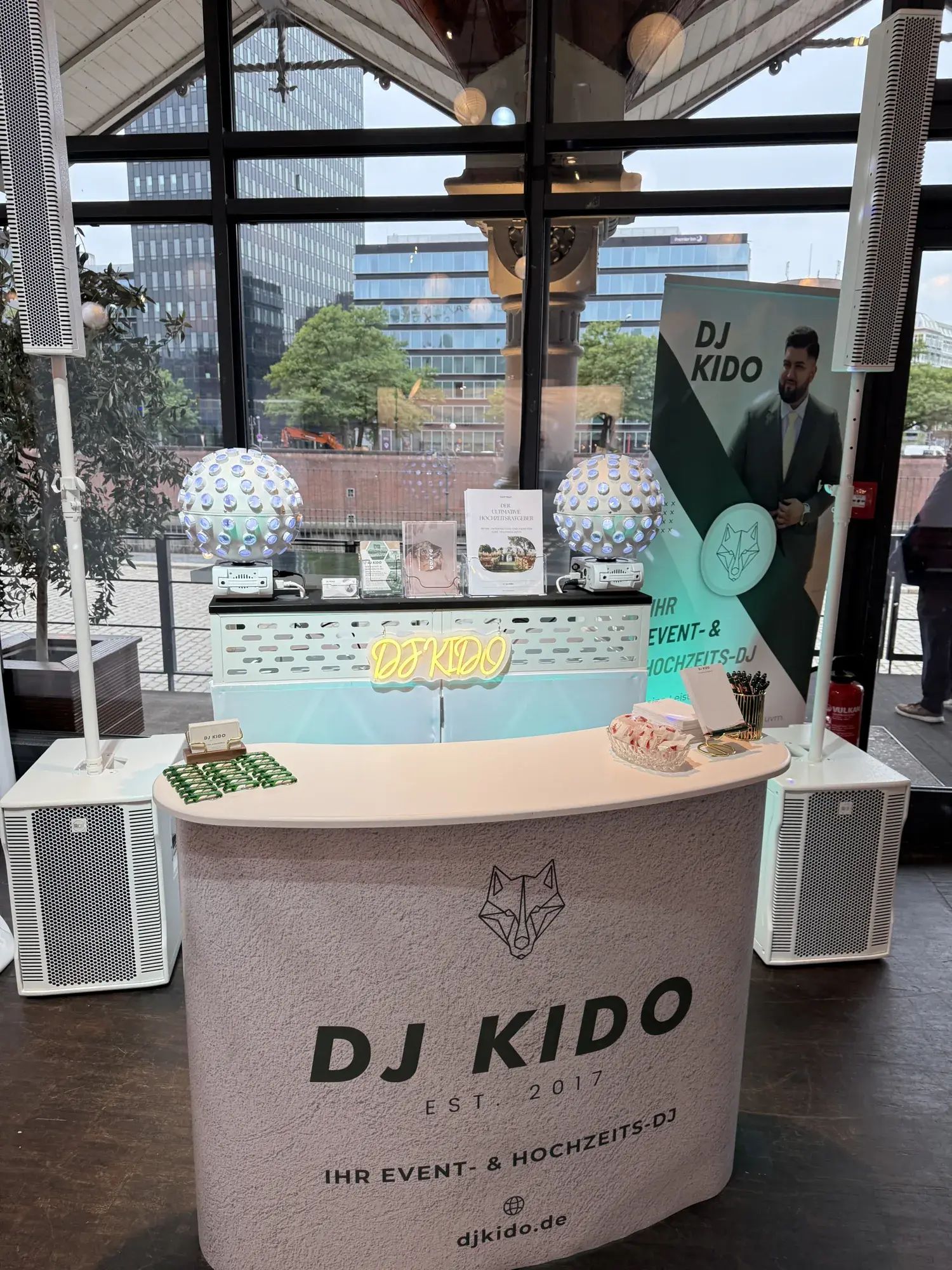 Messestand von DJ Kido auf der BRIDES & GROOMS im Ehemaligen Hauptzollamt: Theke mit Logo, DJ-Booth mit Neon-Schriftzug, zwei Kugel-Lichteffekte, dahinter die Glasfront zum Fleet