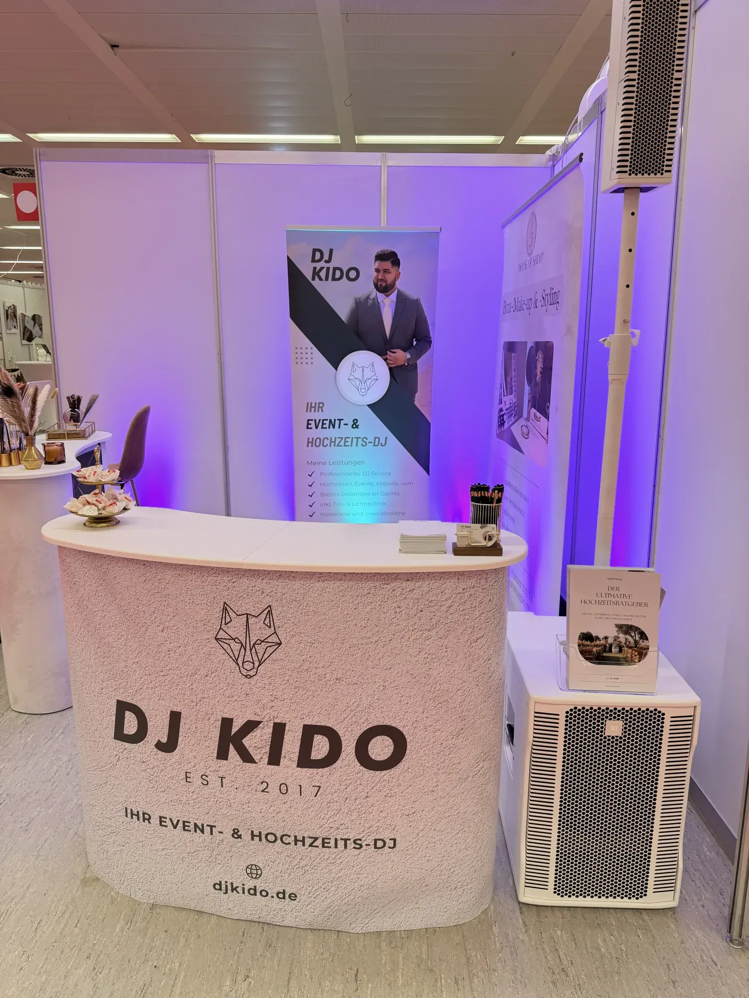 Messestand von DJ Kido auf der TrauDich! Hamburg: Theke mit Logo, Roll-up, Säulenlautsprecher und Subwoofer in violettem Licht