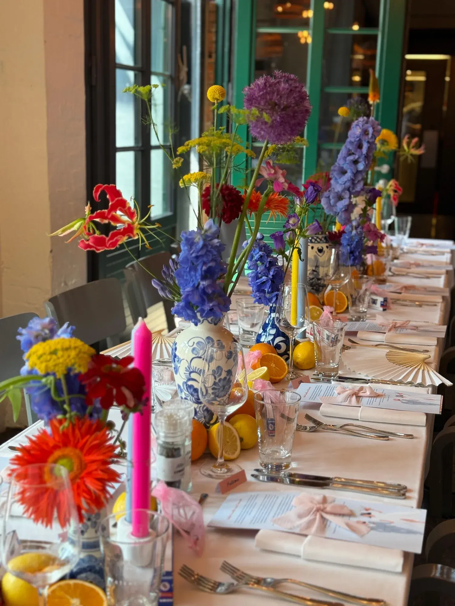 Lange Hochzeitstafel im Catch of the Day mit bunten Blumen in Delfter Vasen, Zitrusfrüchten und grünen Stahlfenstern