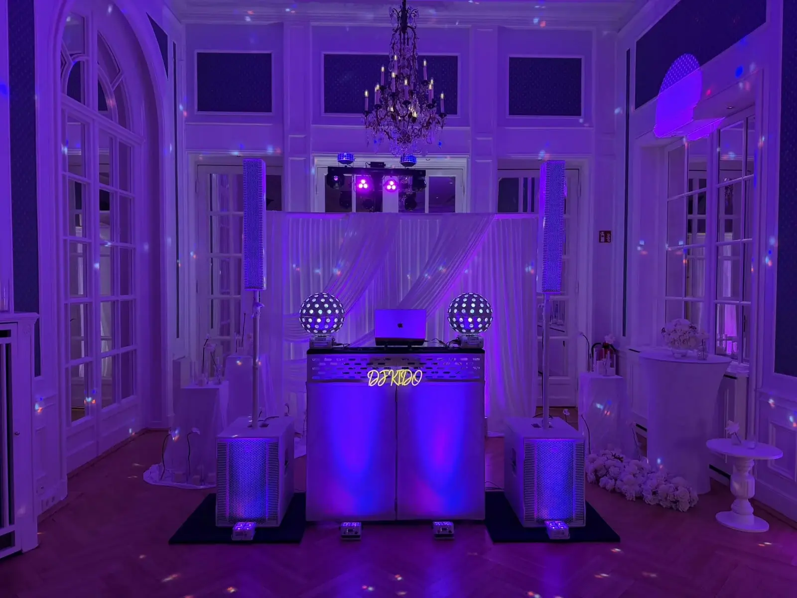 DJ-Setup von DJ Kido im Hotel Atlantic: weiße Booth mit Neon-Schriftzug, Säulenlautsprecher, Subwoofer und Kugel-Lichteffekte vor weißem Vorhang, der Saal in violettem Licht unter dem Kronleuchter