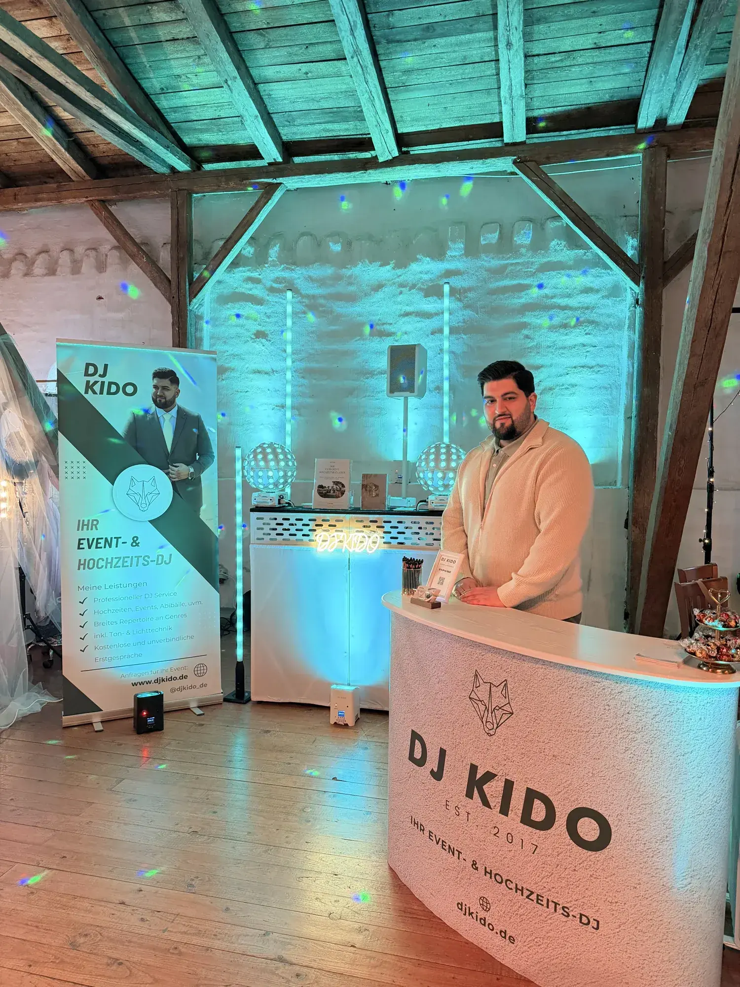 DJ-Kido-Stand auf der Hochzeitsmesse Gut Pronstorf in türkisfarbenem Licht mit Roll-up, Theke und Booth