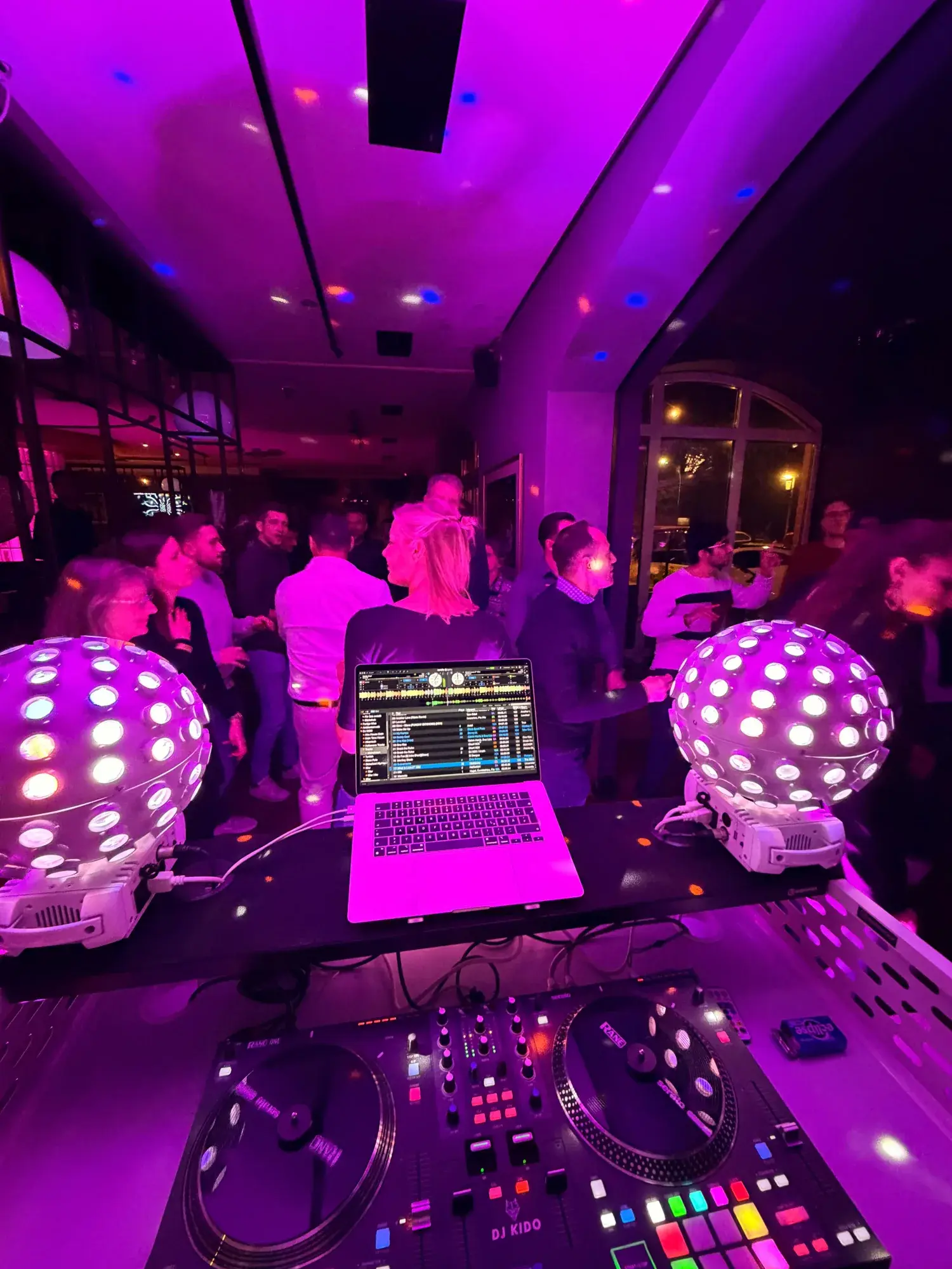 DJ Kido am Pult mit zwei Spiegelkugel-Lichtern, dahinter tanzende Gäste im pink beleuchteten Raum einer Firmenfeier